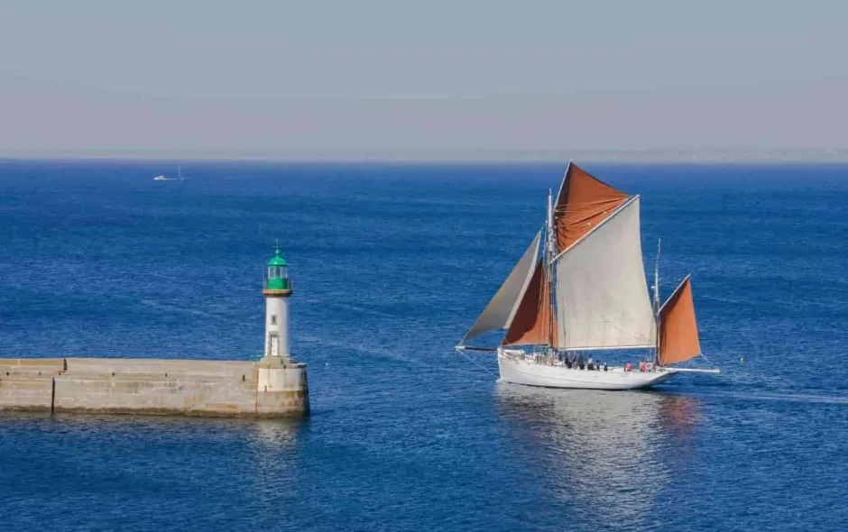 Voilier aux voiles rouges et blanches longeant le phare vert de l’Île de Groix, Bretagne, sous ciel clair et mer calme, ambiance carte postale.