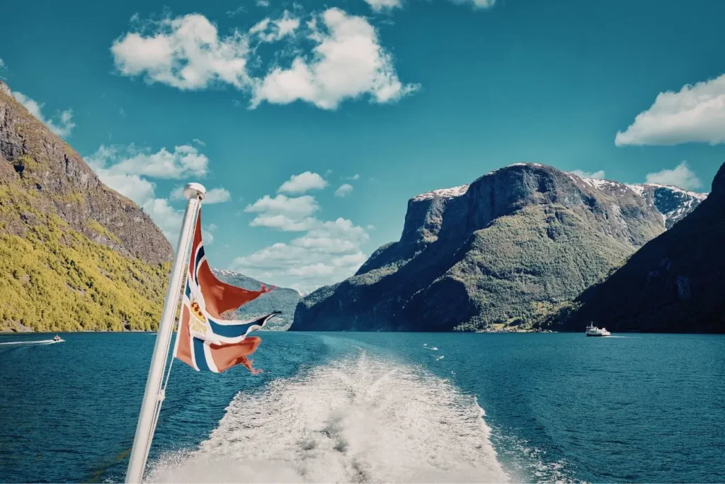Bateau norvégien avec drapeau rouge et bleu avançant sur fjord entouré de montagnes verdoyantes et sommets enneigés, image illustrant la beauté des fjords de Norvège.