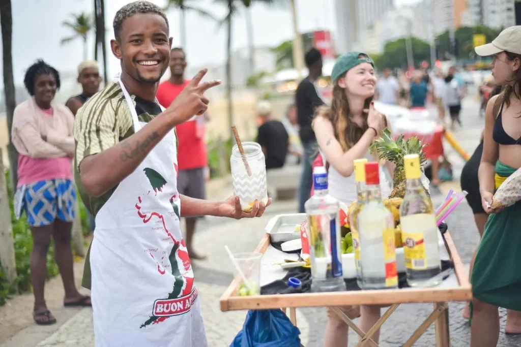 Homme souriant en tablier Buon Appetito tenant un bocal, entouré d’un stand de boissons colorées, capturant l’ambiance festive du carnaval de Rio.