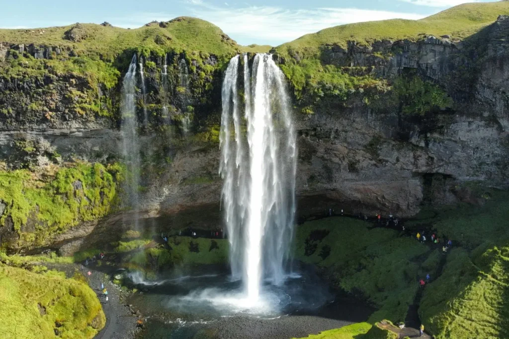 Grande cascade islandaise entourée de verdure et falaises avec visiteurs sur sentier reflétant beauté naturelle et attrait touristique d’un voyage en Islande.