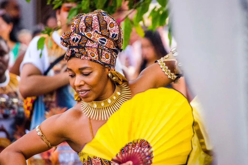 Femme en tenue colorée avec bijoux dorés et éventail, image illustrant la richesse de la culture berbère au Maroc et ses traditions festives.