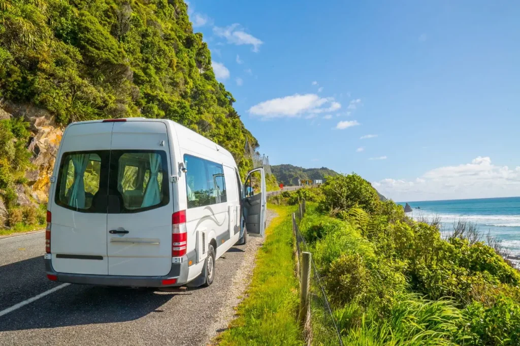 Route côtière bordée de végétation et mer bleue avec van garé, symbole des transports en Nouvelle-Zélande.