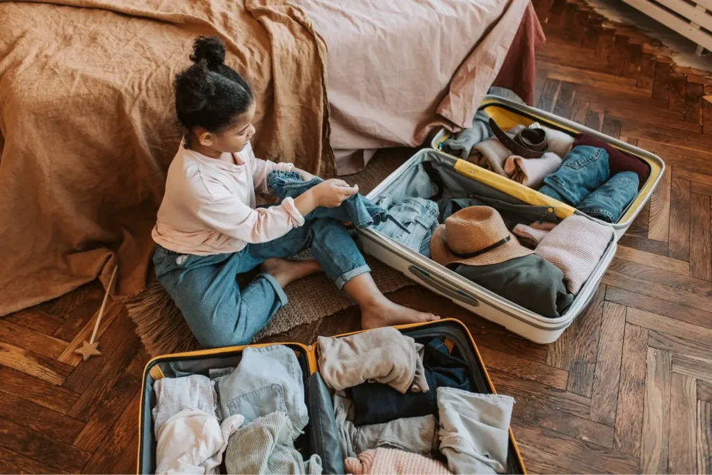 Enfant assis sur le sol rangeant des vêtements dans une valise, image évoquant la préparation d’un séjour chez l’habitant dans un cadre chaleureux.