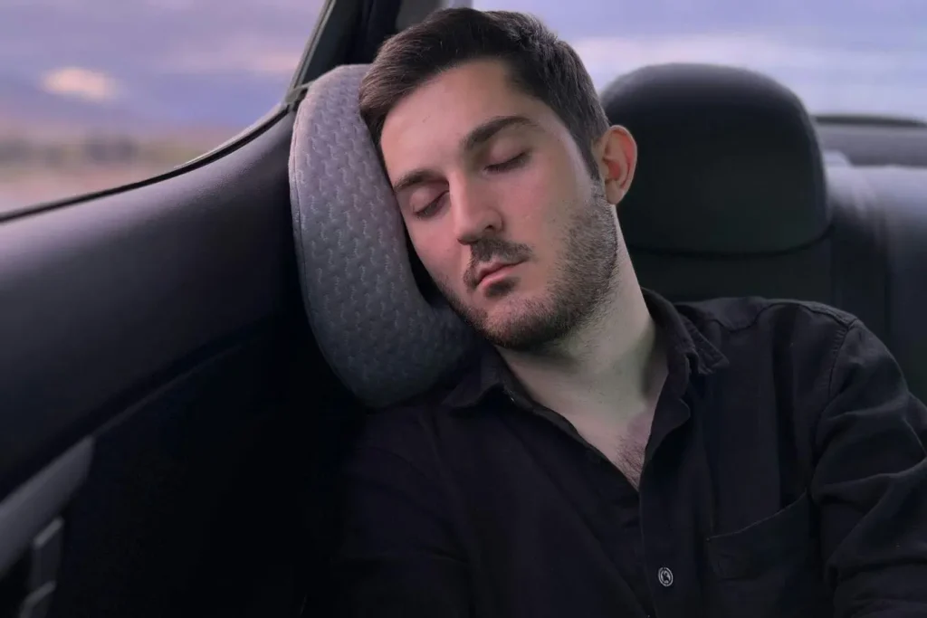 Personne dormant sur oreiller de voyage dans une voiture au crépuscule, image évoquant les astuces pour éviter le jet lag en voyage.