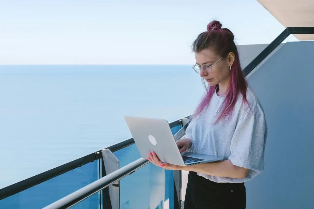 Personne debout sur balcon face à l’océan avec ordinateur portable, image illustrant le nomade numérique et le travail à distance dans un cadre inspirant.