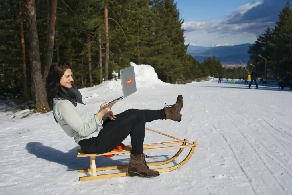 Personne assise sur luge en bois avec ordinateur portable, image illustrant le nomade numérique et le travail à distance en pleine nature enneigée.