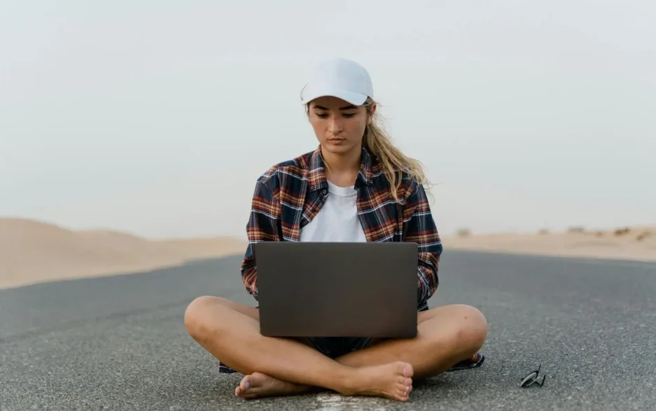 Personne assise en tailleur sur route désertique avec ordinateur portable, image illustrant le nomade numérique et le travail à distance depuis n’importe où.