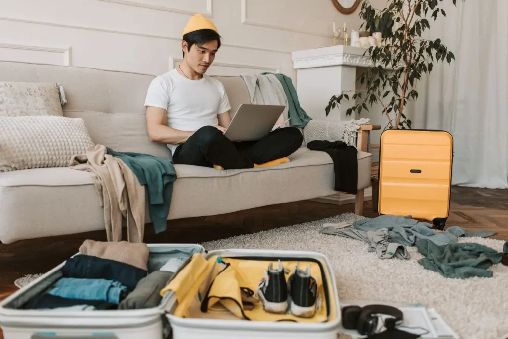 Personne assise sur canapé avec ordinateur portable et valises ouvertes, image évoquant la préparation d’un séjour chez l’habitant dans un cadre convivial.
