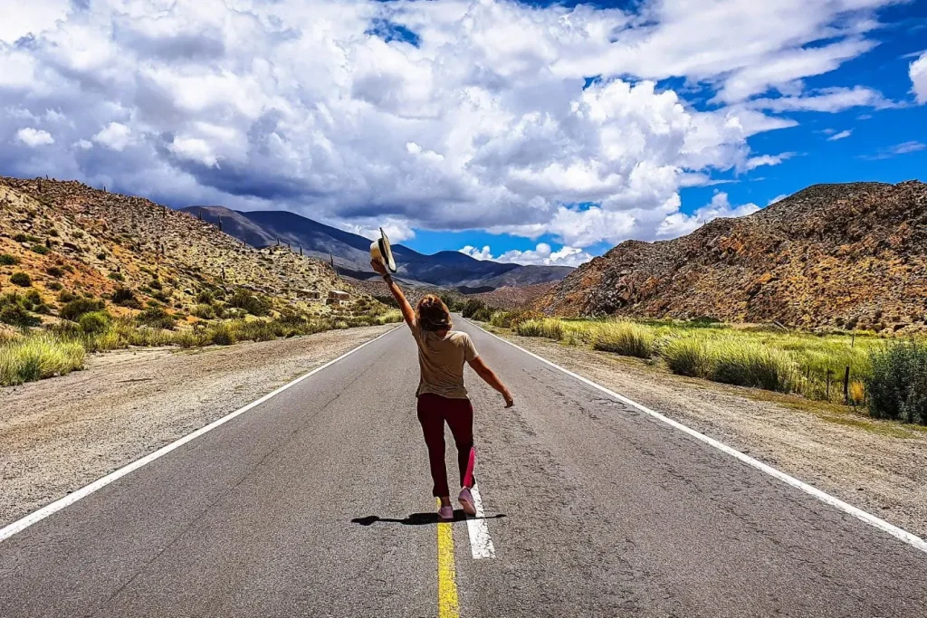 Voyageur solitaire avançant sur route vide entre collines arides, symbole d’indépendance et de découverte personnelle.