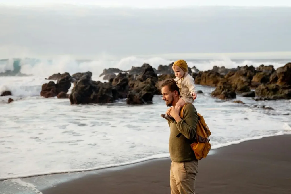 Adulte portant bébé sur épaules en marchant au bord de mer, image représentant comment voyager avec un bébé de moins de 2 ans en toute sérénité.