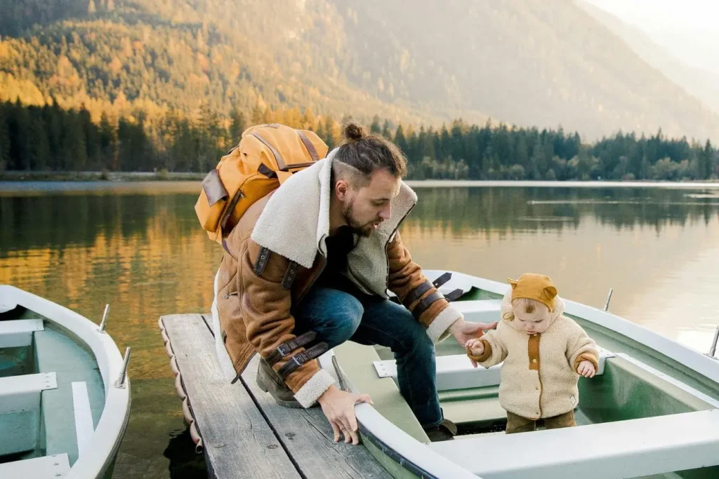 Adulte en veste brune tenant la main d’un bébé sur un bateau au bord d’un lac, image représentant comment voyager avec un bébé de moins de 2 ans en nature.