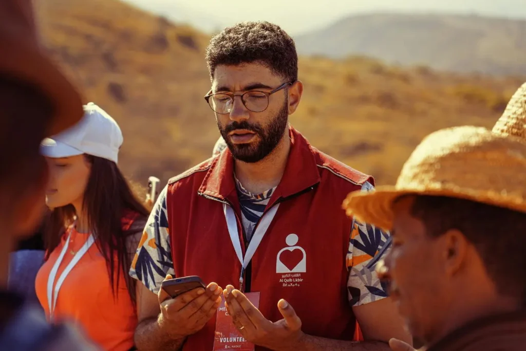 Un bénévole barbu portant un gilet rouge consulte son téléphone lors d'un voyage humanitaire en plein air entouré de membres de son équipe solidaire.