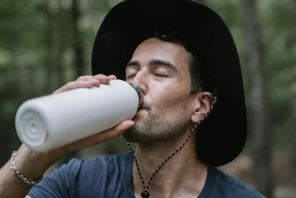 Voyageur portant chapeau sombre buvant à une gourde réutilisable en forêt, illustrant l’importance de l’eau sûre pour éviter la tourista en voyage.