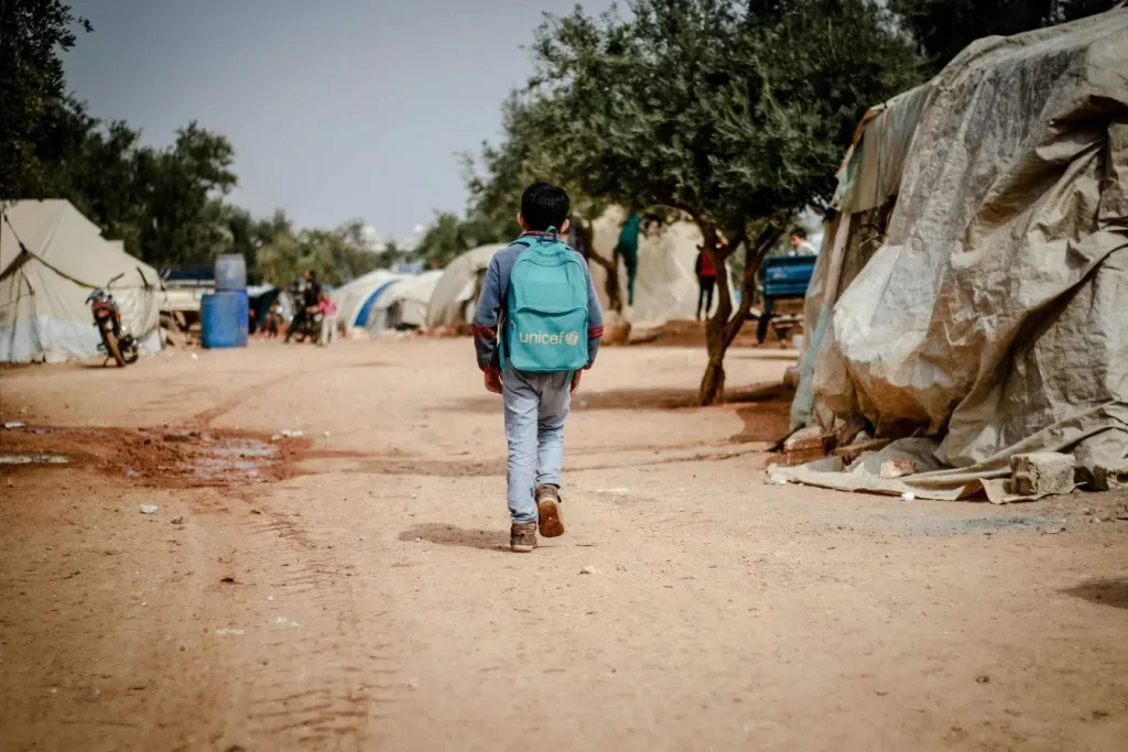 Un enfant marche de dos dans un camp de tentes poussiéreux, portant un sac à dos bleu clair siglé UNICEF, illustrant l'impact concret de l'aide lors d'un voyage humanitaire.