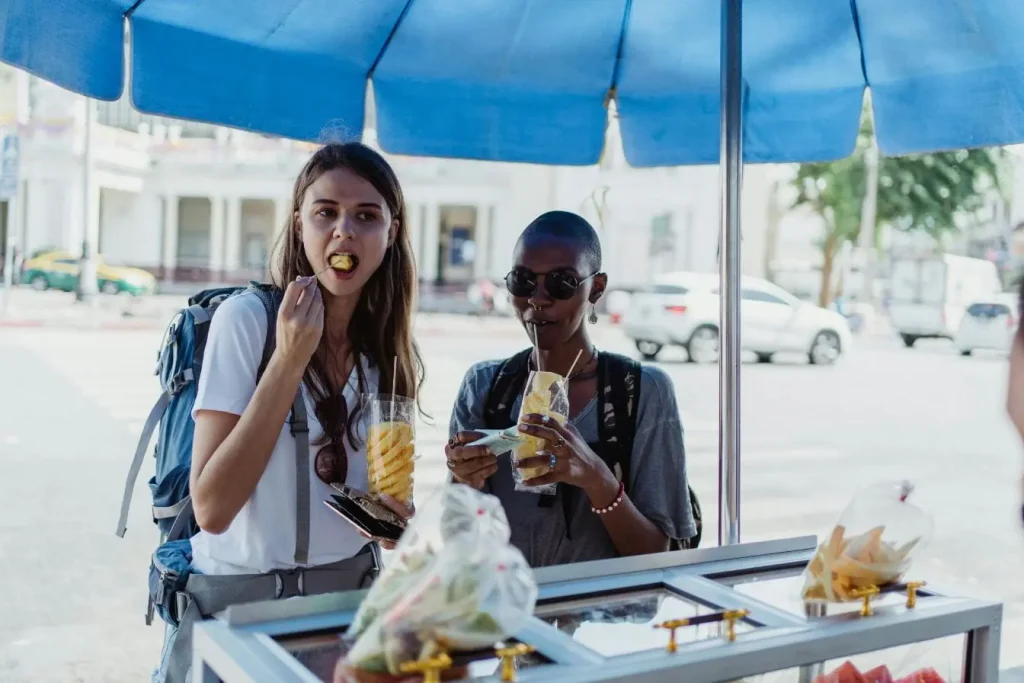 Deux voyageurs sous un parapluie bleu dégustent des fruits frais en gobelets, illustrant l’importance de choisir des aliments sûrs pour éviter la tourista en voyage.