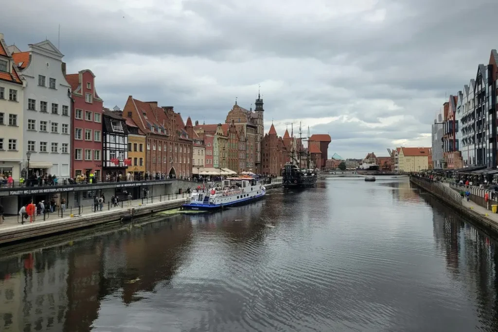 Vue sur les quais de Gdańsk, bâtiments colorés bordant la rivière avec voilier ancien et bateau moderne amarrés.