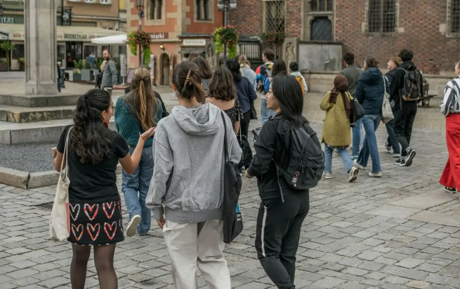 Groupe de visiteurs explorant Wrocław, marchant sur rue pavée entourée de bâtiments historiques ornés de fleurs et boutiques locales.