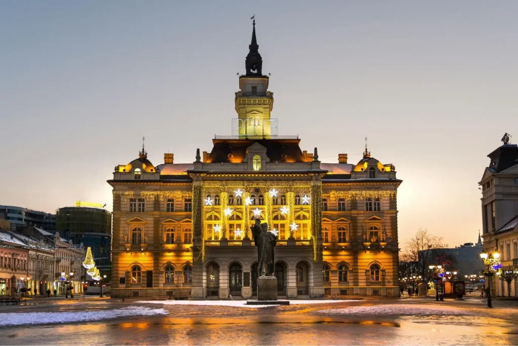 Place enneigée devant bâtiment illuminé et statue, reflet des incontournables festifs à découvrir en visitant Novi Sad.