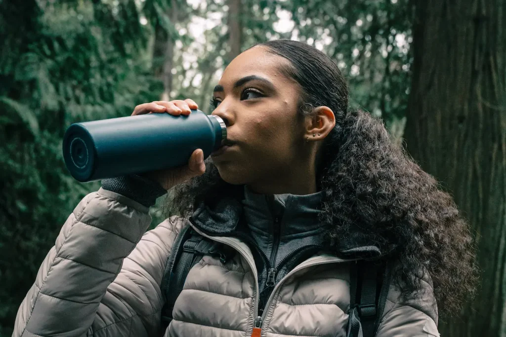 Personne en veste grise buvant à une gourde bleue en forêt, reflet du moment de pause et d’hydratation lors d’un repas en randonnée.